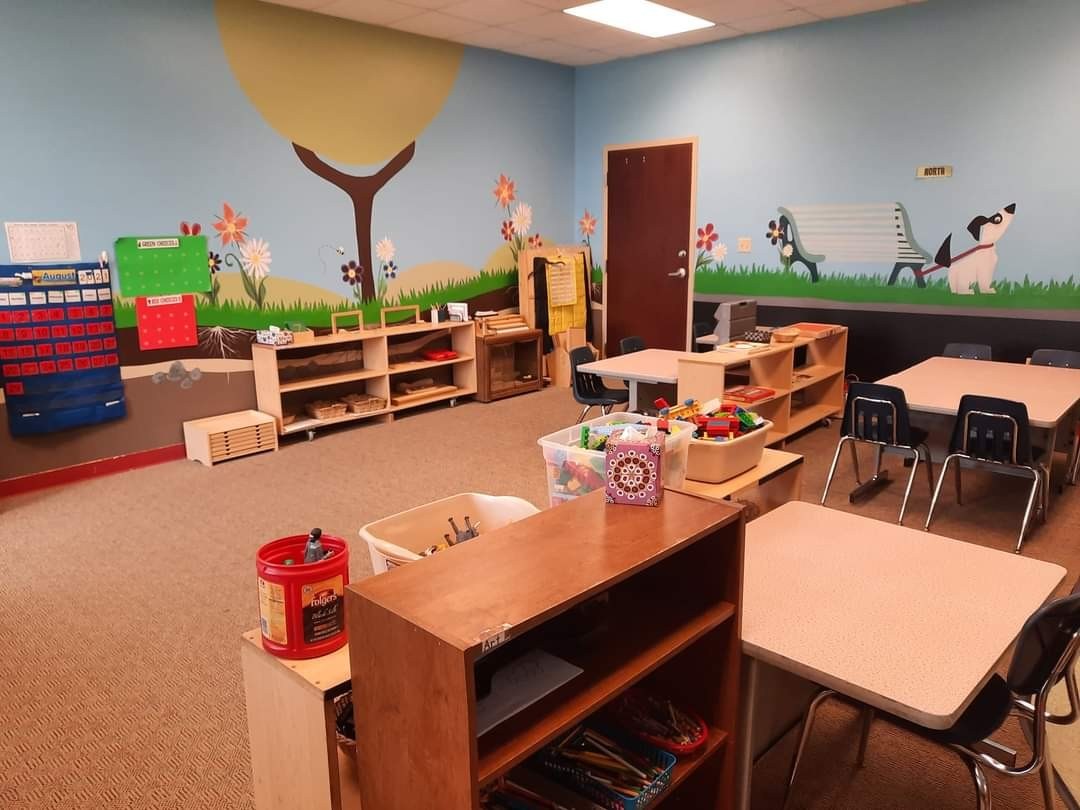 The classroom, with various tables, shelves, and a door.