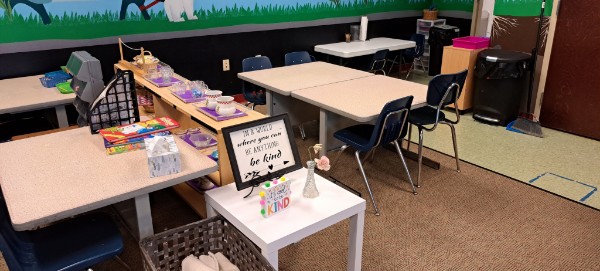 A close-up of a couple tables. A sign on one of them reads: In a world where you can be anything, be kind.